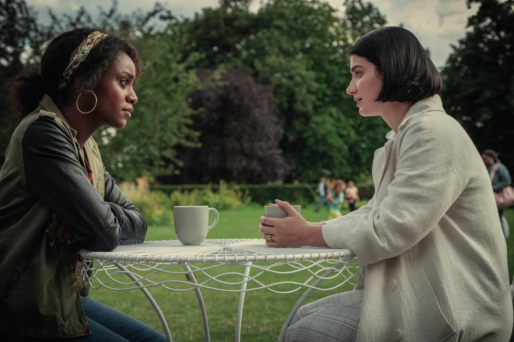 Eve Hewson and Simona Brown sit at a coffee table outdoors in character for Behind Her Eyes