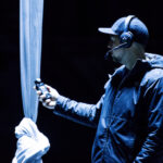 A man with a cap standing next to an aerial silk. He holds a control