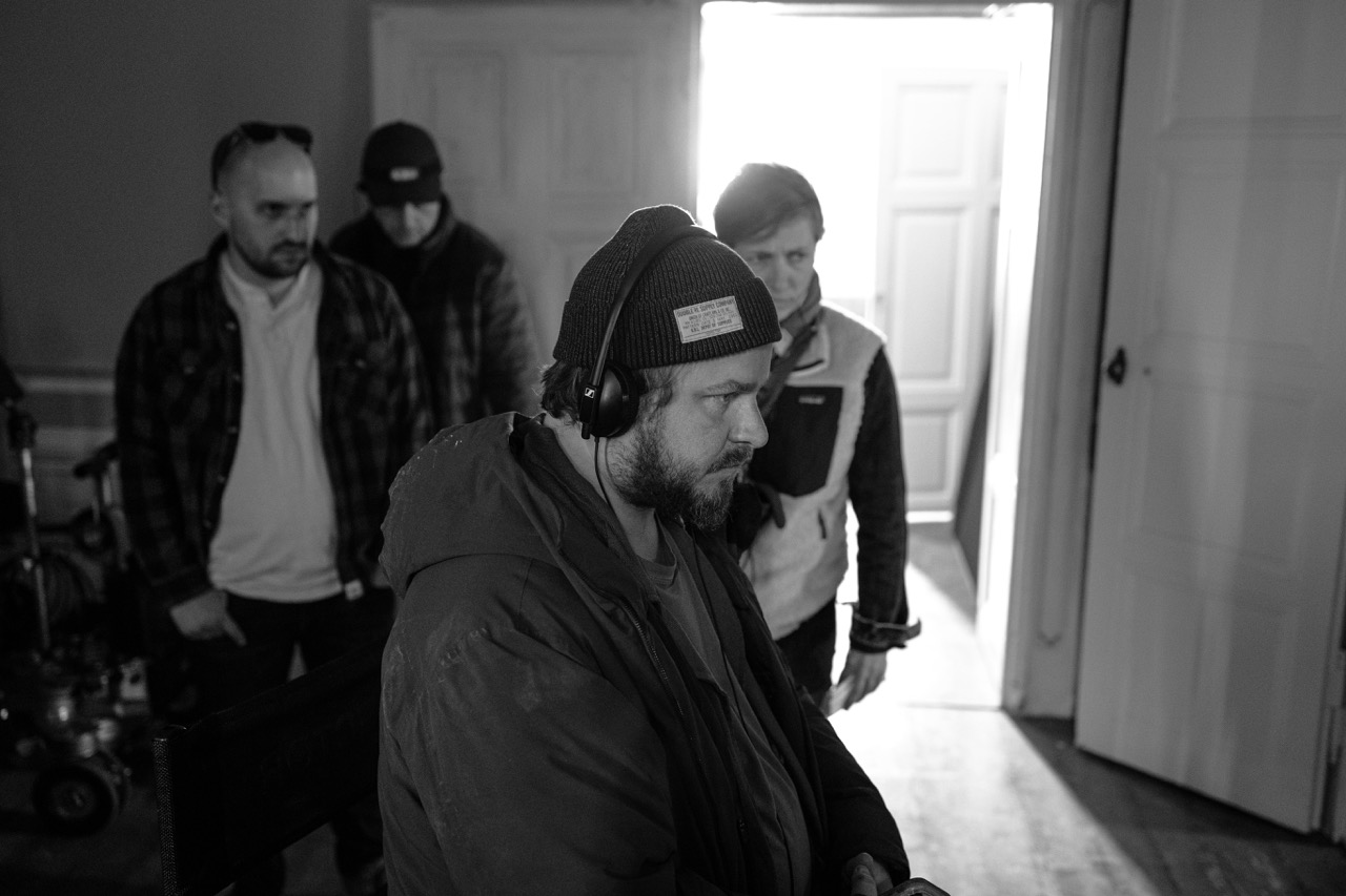 A director in a beanie and headphones focuses intently on a monitor, while crew members stand behind him in a dimly lit room