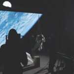 A person in a space suit dangling from a harness in a dark room while being filmed in front of a blue background