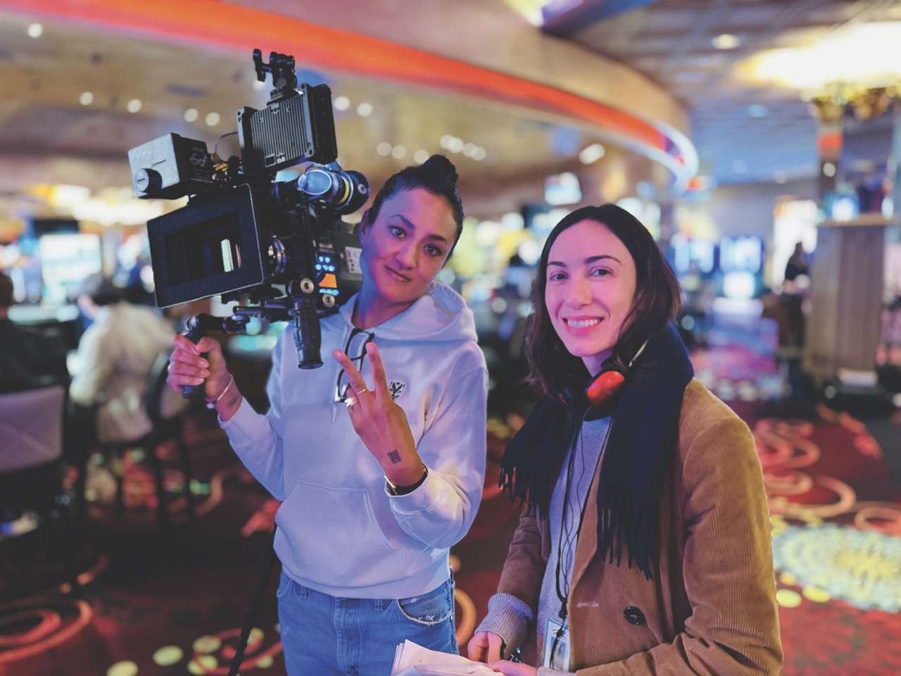 Two people smiling. One holds a camera on their shoulder and the other has headphones around their neck