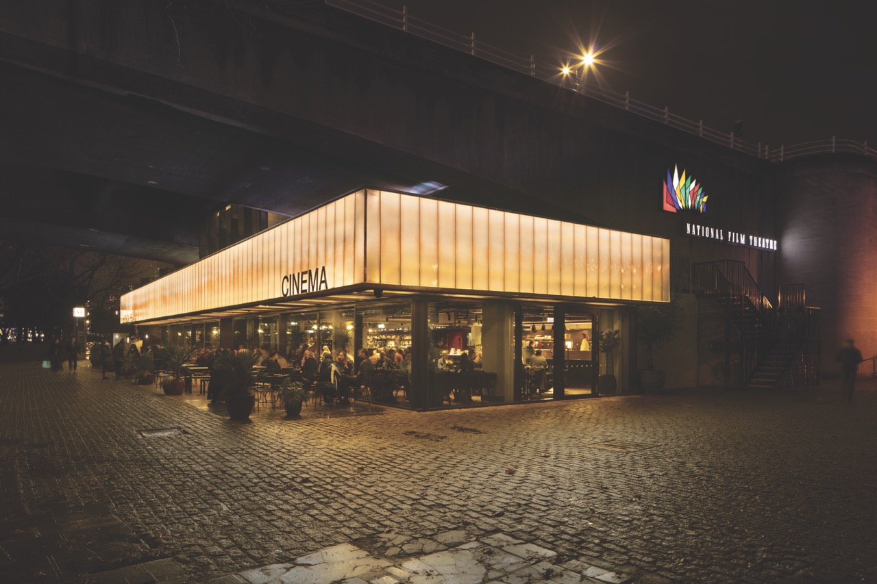 The outside of a low cinema with an illuminated roof and large glass windows. The cinema is under a bridge