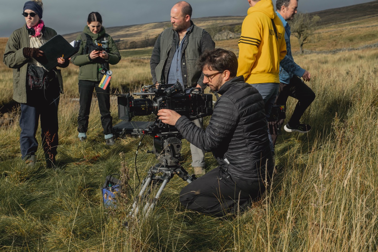 Man in a black coat kneels in a field with a large camera while people stand behind