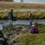 Three children sitting in a field near a river with a sound boom to the right of the shot