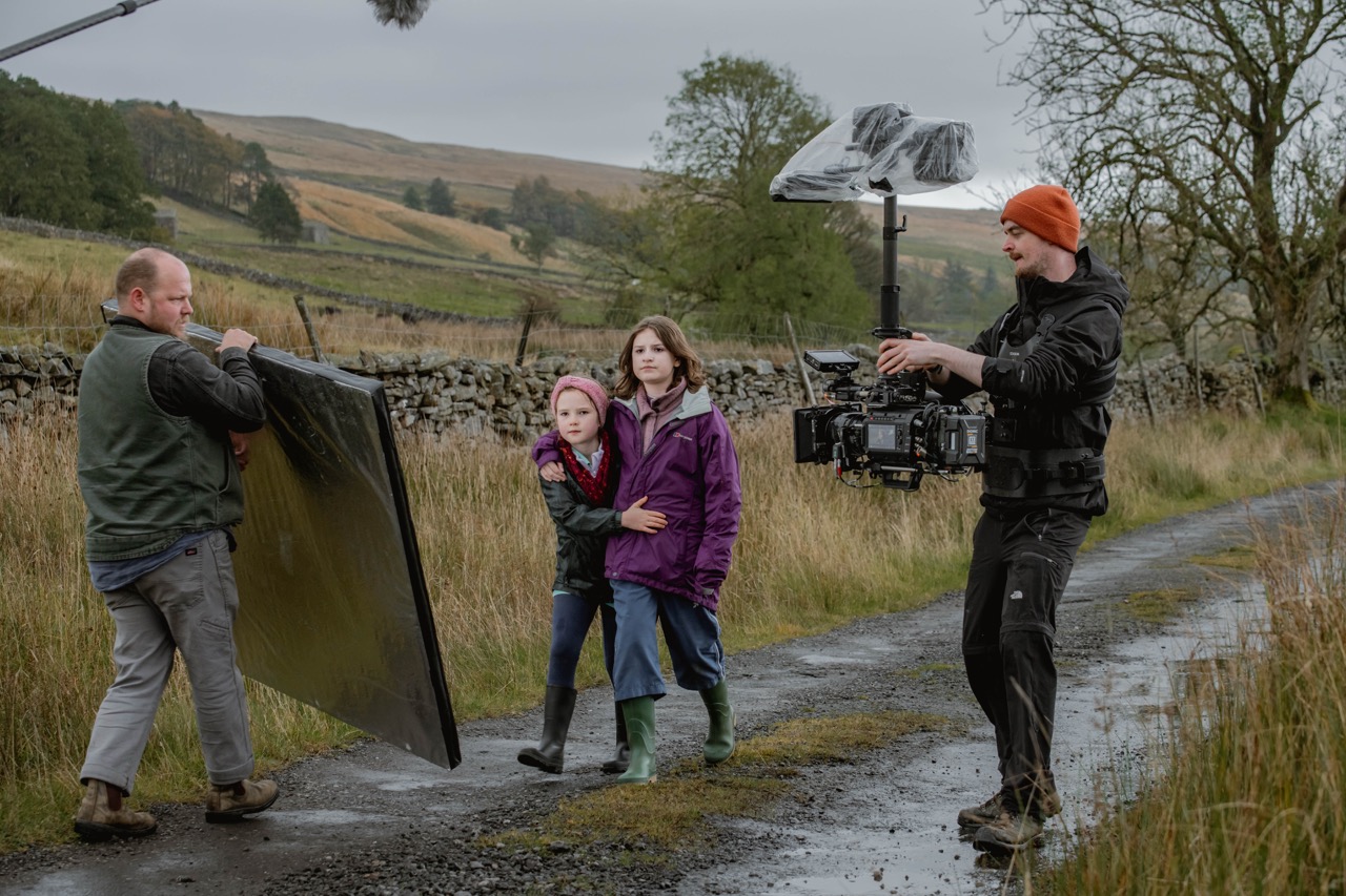 Two children walk down a path in the countryside while being filmed by two men. One man holds a camera and the other holds a reflective board.