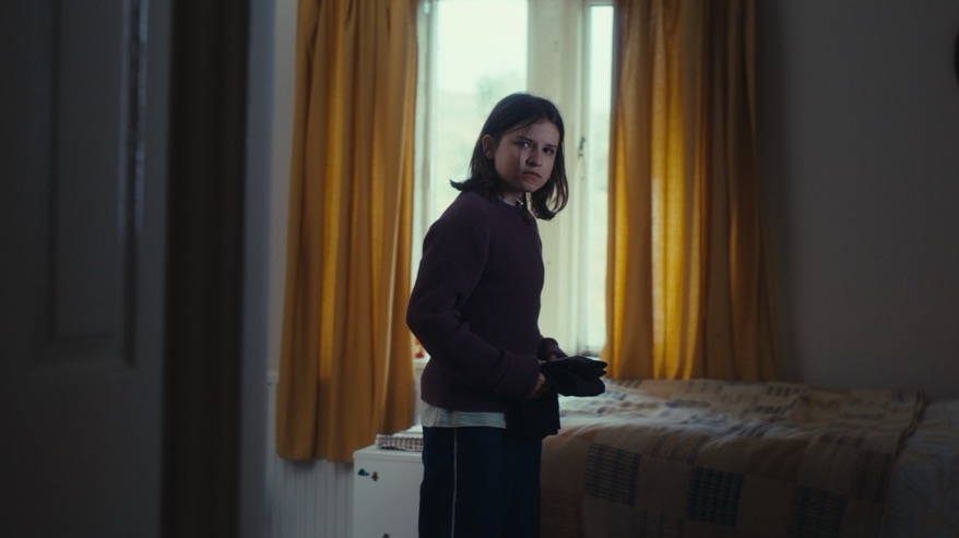 A girl stands in front of a window with yellow curtains