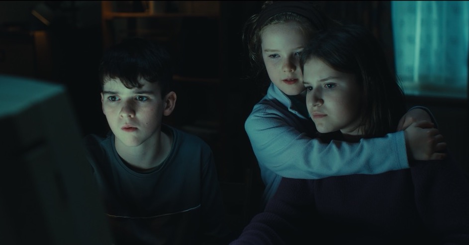 Three children sit in a dark room. Two of them are hugging tightly.