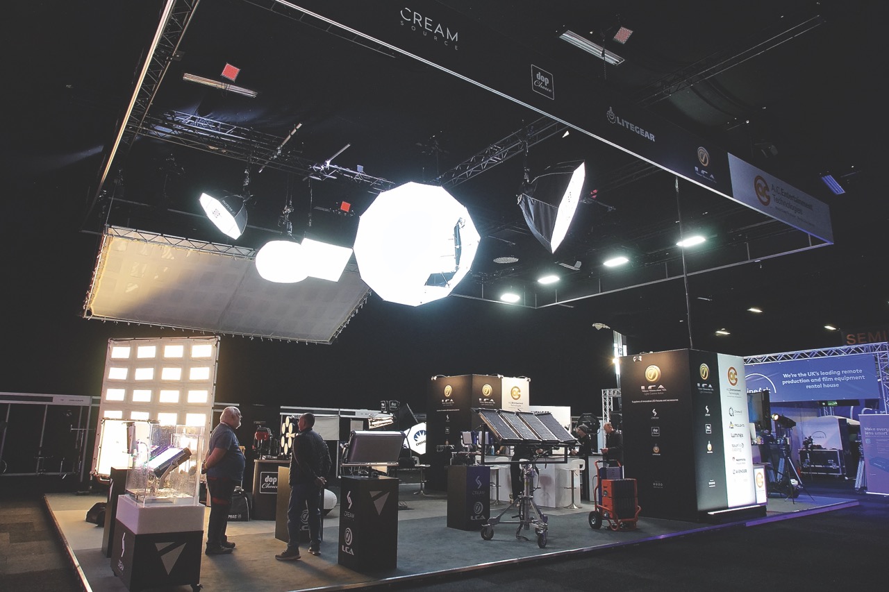 A group of people standing in a room in an exhibition where there are stands set up and large lights hanging from the ceiling