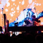 A person playing a music on stage with an orange and blue background. Image Genevieve Reeves & Seb Gardner