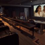 A dark cinema room with tiered seating and a large screen showing a man and woman onscreen