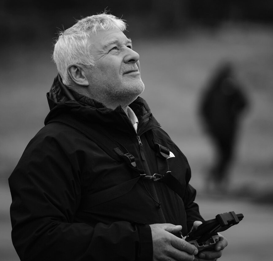 Black-and-white photo of a man in a coat holding a remote looking at the sky