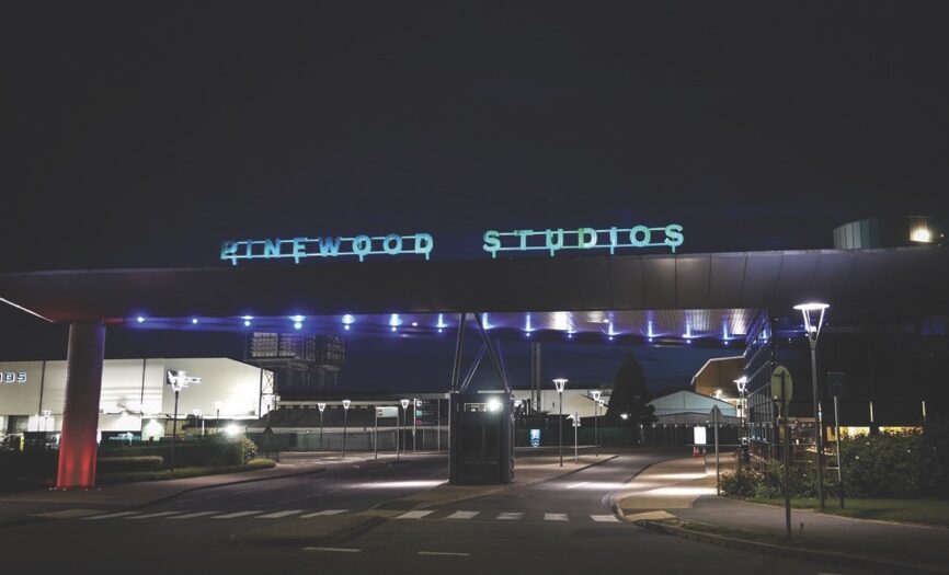 The outside of a building at nighttime. The building says "Pinewood Studios" in blue glowing writing.