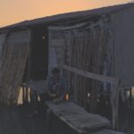 A person sitting in front of a wooden plank house in the ocean at sunset