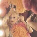 A showgirl standing in front of a light-ringed mirror putting on a feathered head piece