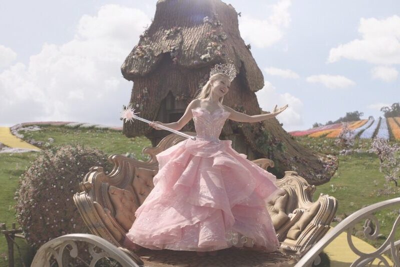 A woman wearing a large pink dress with a wand. She stands on a carriage in front of a field and wooden house