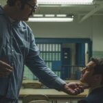 A man in prison clothing stands and stroke the chin of a seated man wearing a suit