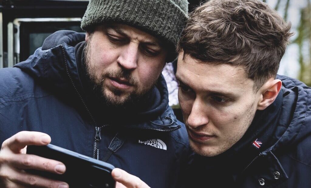 Two men stand close to each other outside, looking at a phone screen