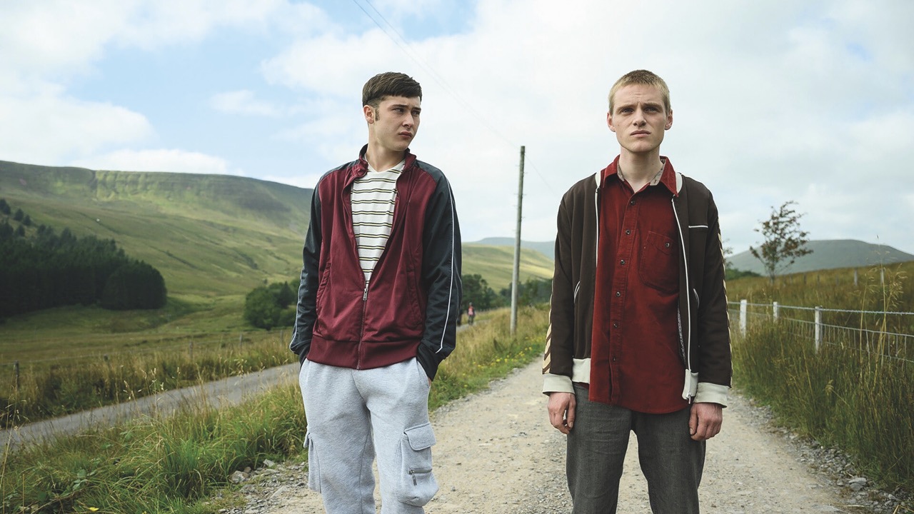 Two young men stand on a path in the countryside. They look angry