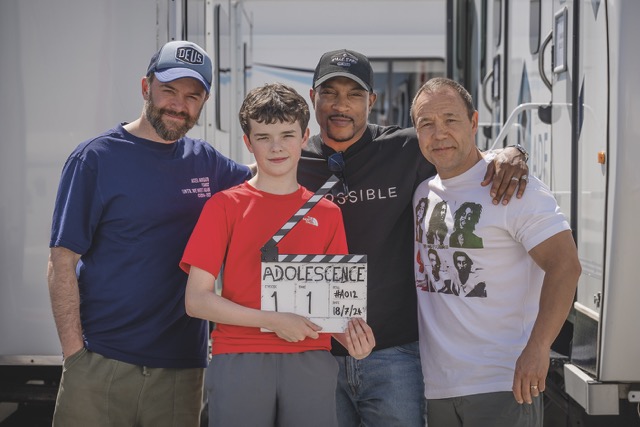 A boy in a red shirt stands with three men. The boy holds a movie slate