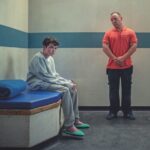 A young boy wearing a grey tracksuit sitting in a prison holding cell while a man with an orange shirt stands nearby