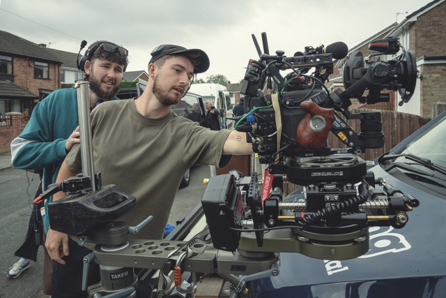 A man operates a large camera outside while another man supports him from behind