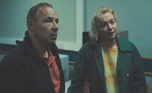 A man and a woman stand wearing coats in a grey room. They look concerned