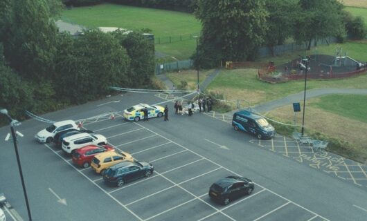 An aerial shot of a car park next to a field with a police car and police tape in one corner
