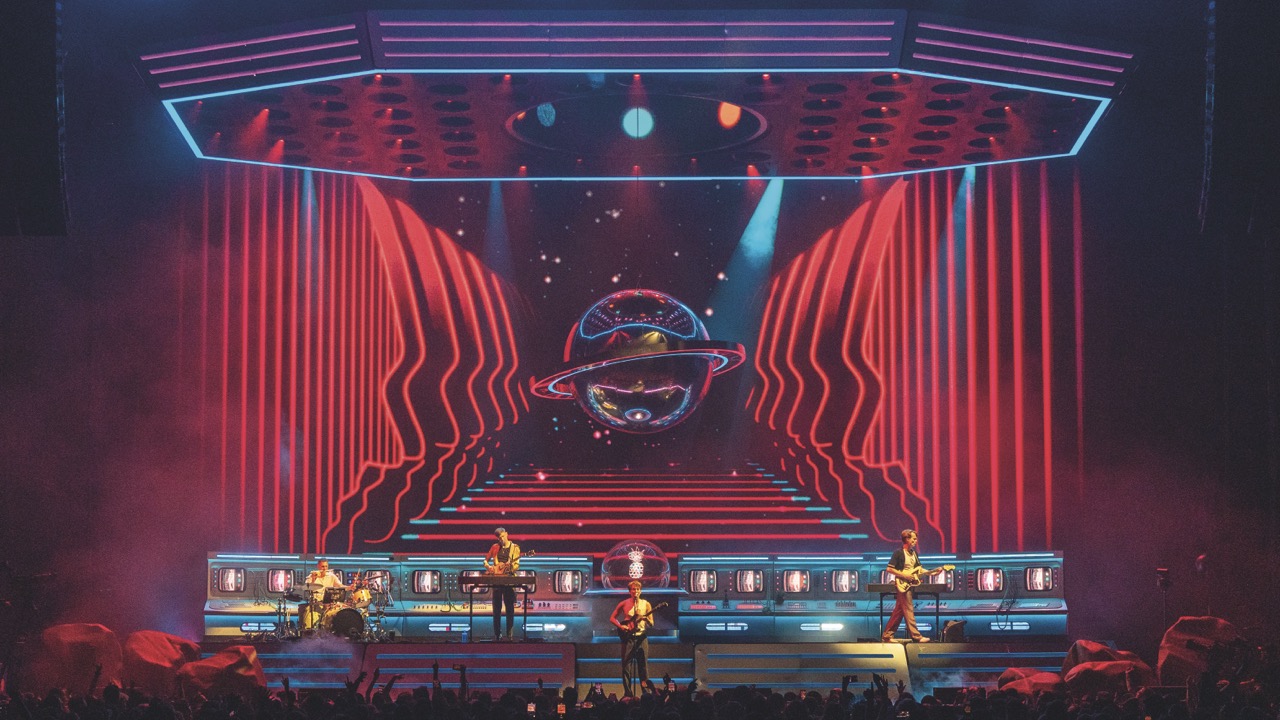 A band on a stage with a large LED screen behind showing red animations and a planet