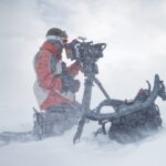 A person wearing red snow gear and skis. They are kneeling in a snow storm, looking through a camera
