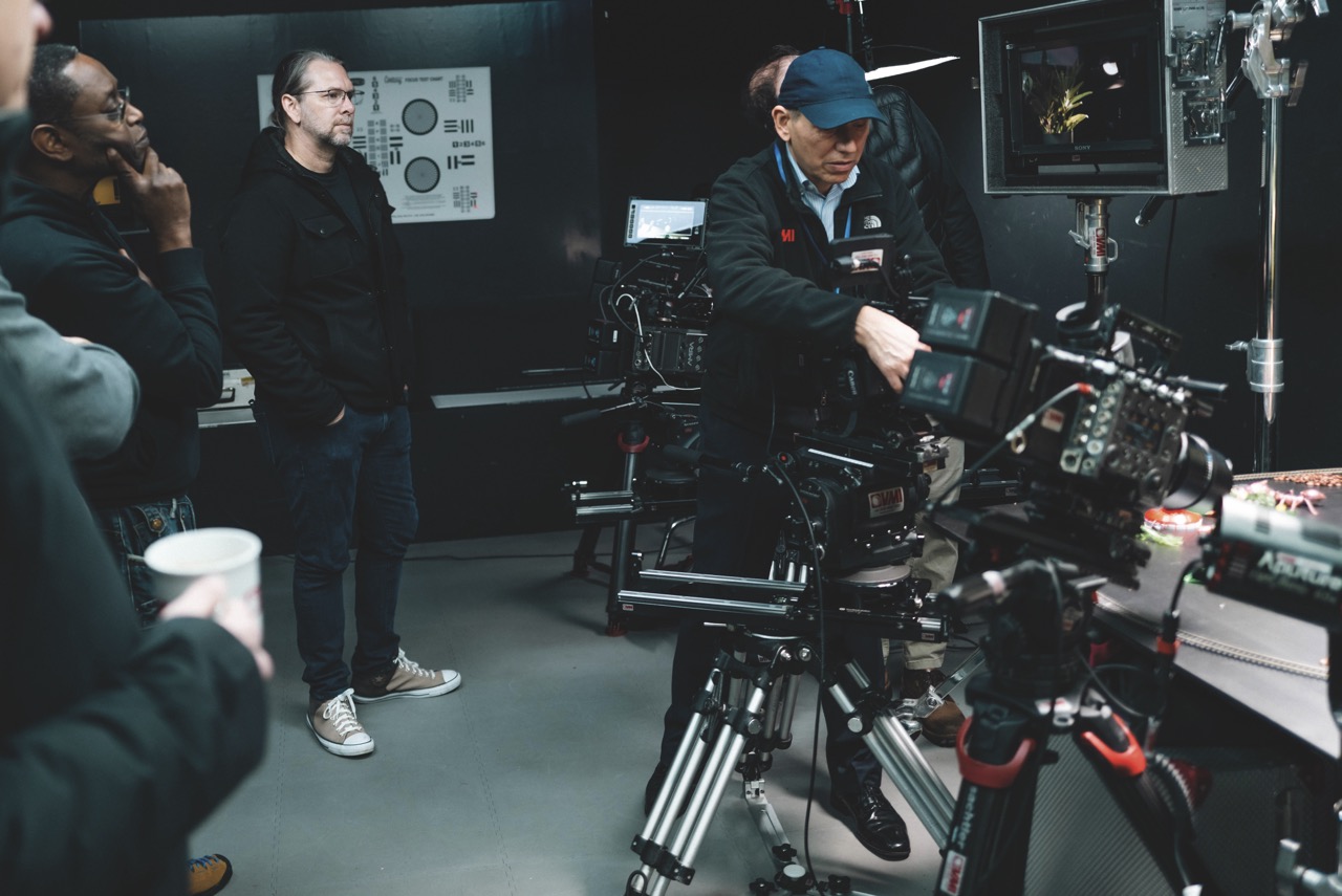 A man wearing a cap operates a camera while a group stands around and watches