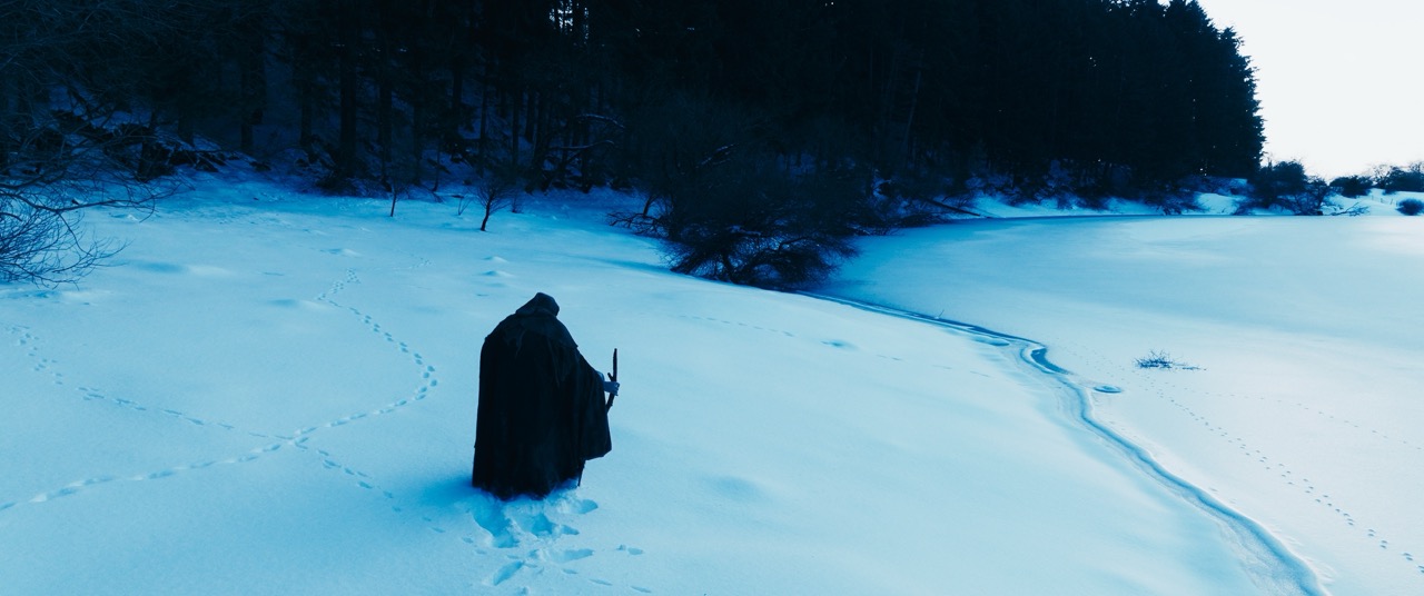 A person in a thick black cloak journeys across a snowy terrain