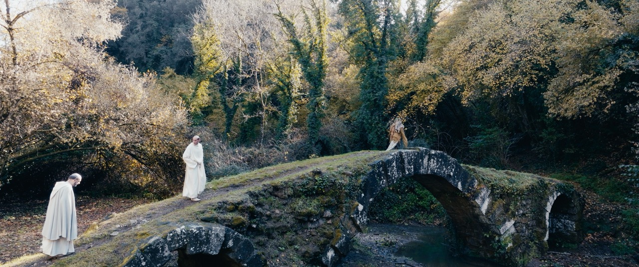 Two men in white robes cross a grassy bridge in a forest