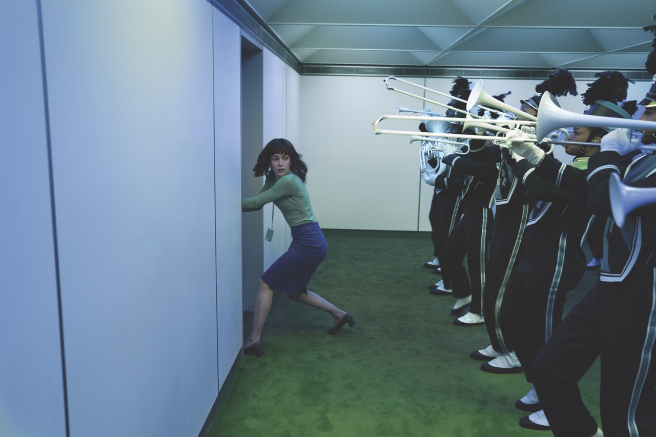 A woman trying to get through a door in a room with green carpet. A marching band stands in a row behind her