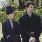 An older woman walking with a younger man near a hedge. They are dressed in period dress