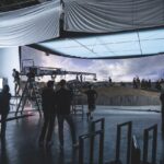 A filming set with a large LED screen showing a background. A team stands around monitoring and filming the scene