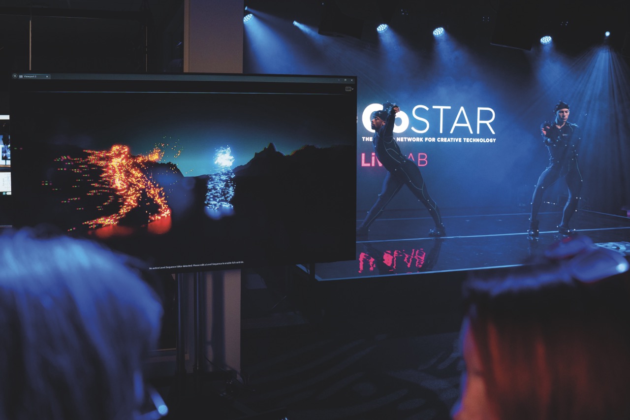 Performers dancing on a dark stage with a monitor converting the image in front of an audience