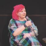 A woman with bright red hair holding a microphone and presenting on a stage