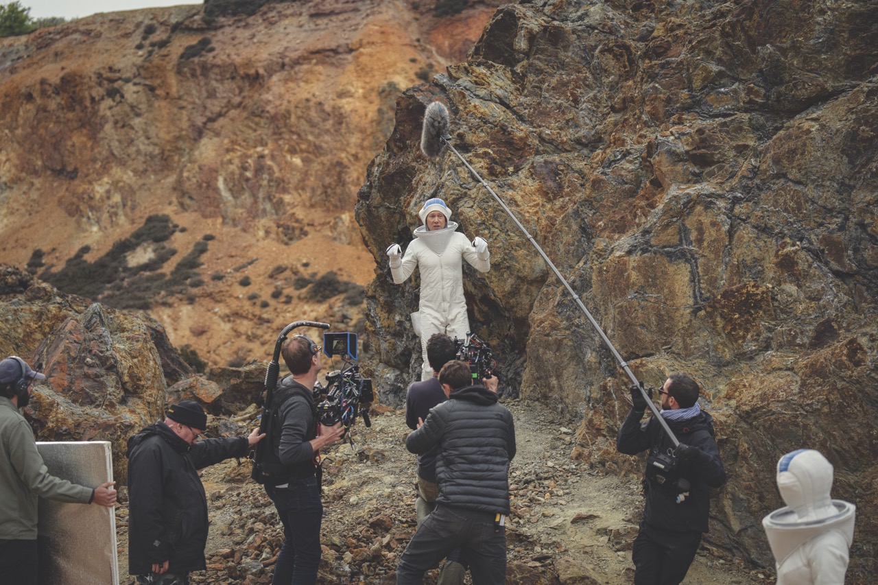 An actor wearing a white space suit being filmed by a crew with a large sound boom