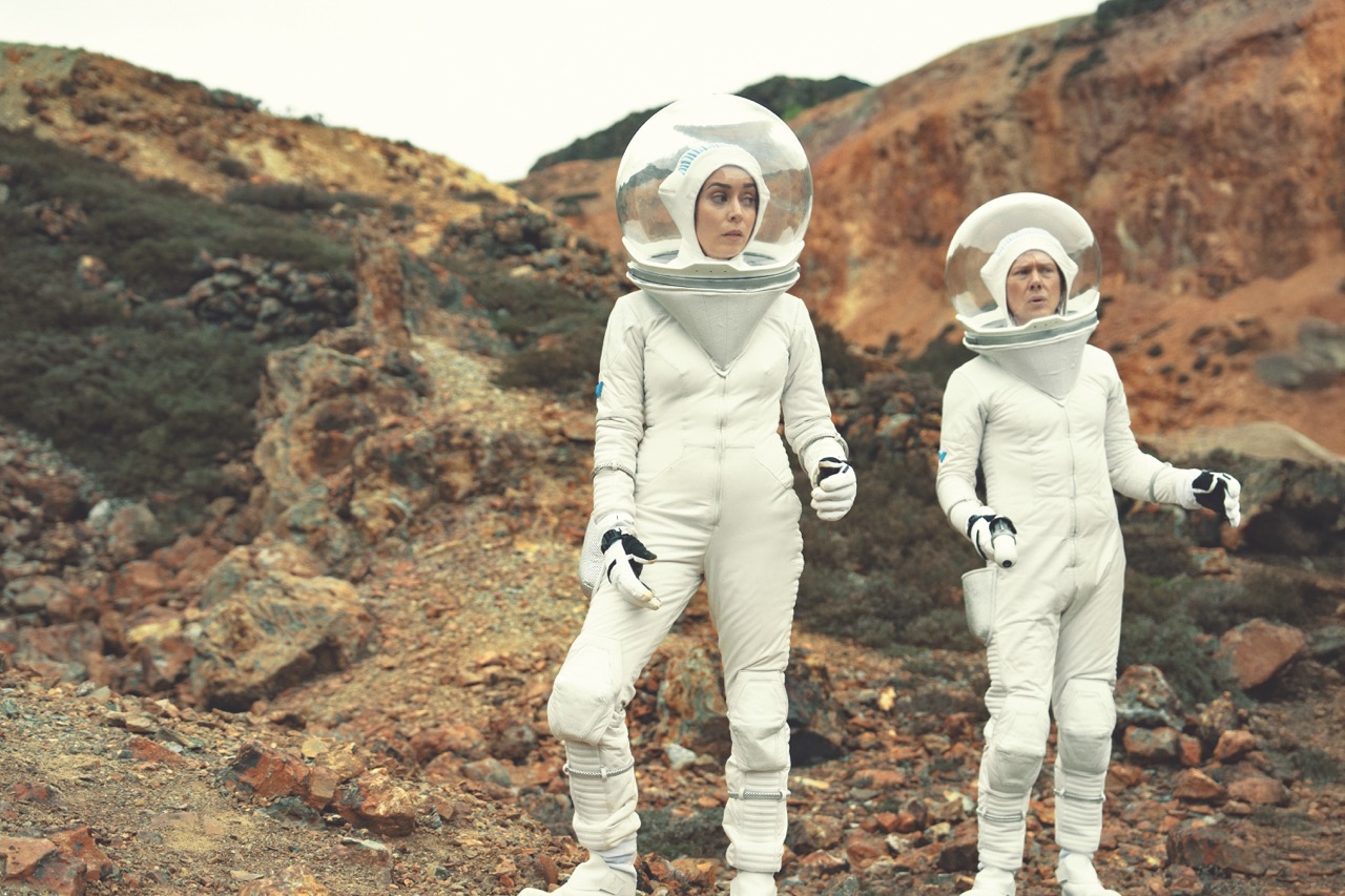 Two people standing in white space suits, standing in a barren terrain