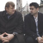 Two men sit with their hands clasped in their laps. They wear suits and sit outside looking at each other