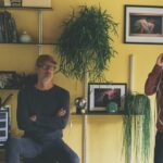 Two men standing in a yellow room. One sits on a stool while another talks into a headphone
