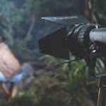 A professional lighting device set up in a wooded area. A person sits near a camp fire in the blurred background
