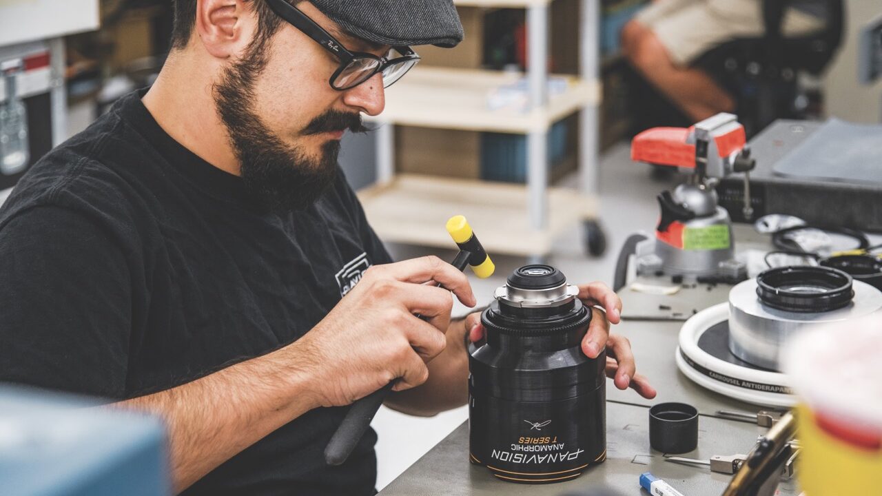 A man with glasses and a flat cap working on a camera lens with a small hammer