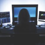 A person sitting in silhouette facing three computer screens filled with numbers and graphs