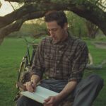 A man sitting on the grass under a tree, writing in a diary. There is a bike standing behind him