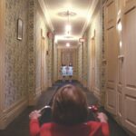 A person rides a bike along a hallway towards two twins dressed in blue dresses