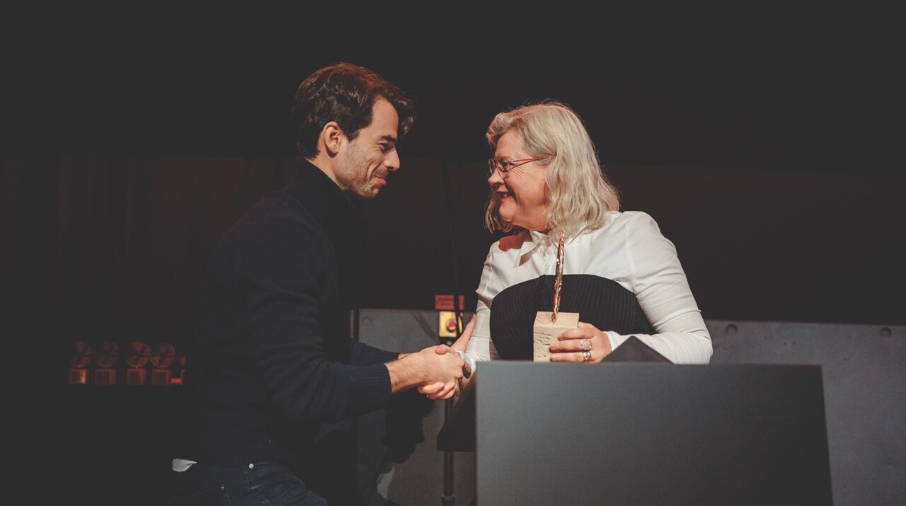 A man and woman exchange a handshake and award on a stage