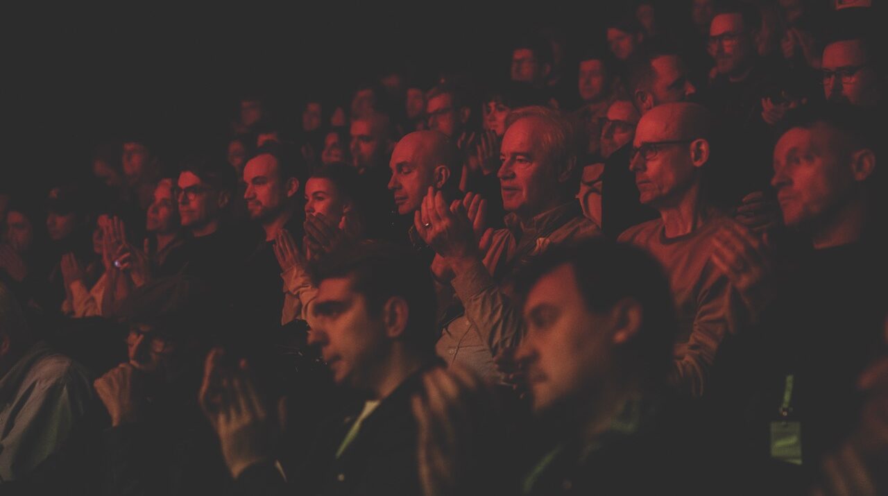 A crowd sitting on tiered seating in dark red lighting