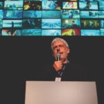 A man standing on stage with a microphone while multiple pictures appear on the projector behind him
