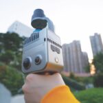A hand with an orange sleeve holds a silver scanning device in a park in a city